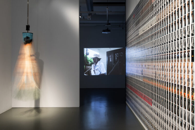 A kinetic sculpture blurs from motion beside a large projection of a mail carrier, with an adjacent wall covered in a dense grid of small photographs.