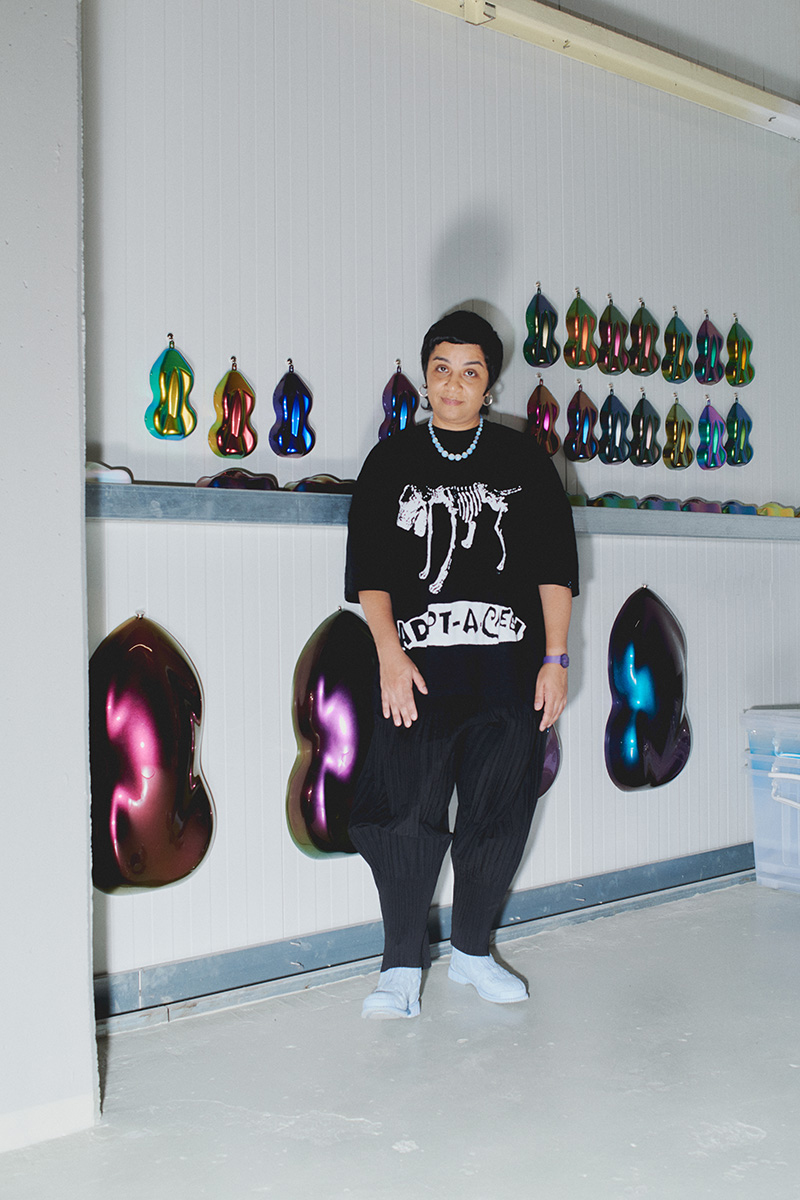Monira Al Qadiri posing in front of a wall with iridescent plastic wall works hanging on it