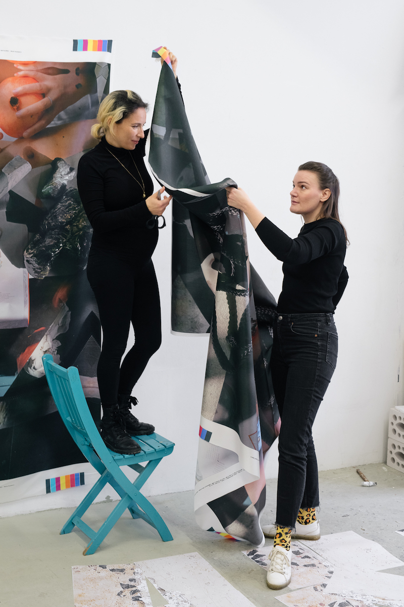 two artists hang a textile print on the wall, one standing atop a blue folding chair